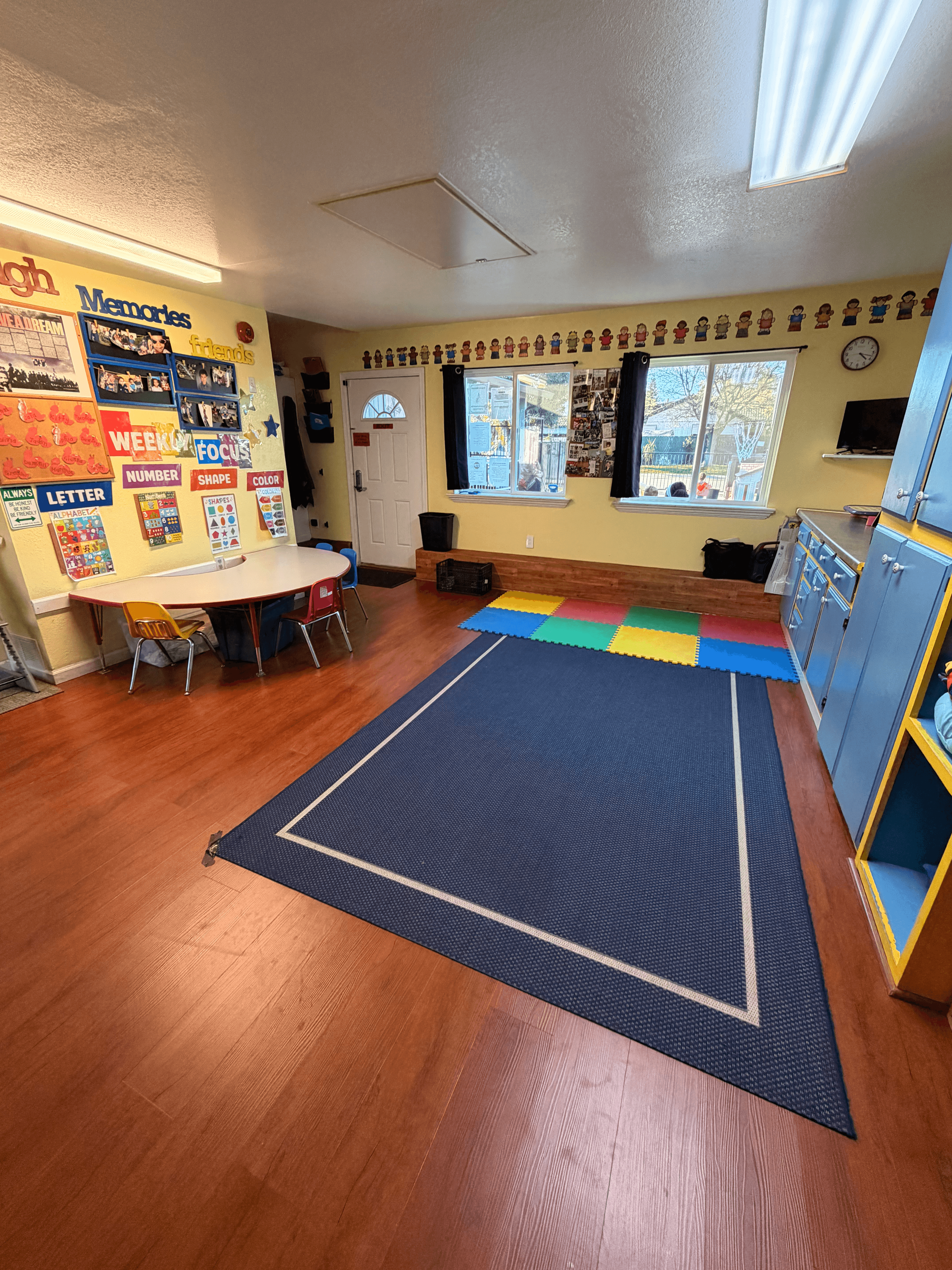Preschool classroom featuring a large blue rug, wood floors, and walls decorated with educational posters.
